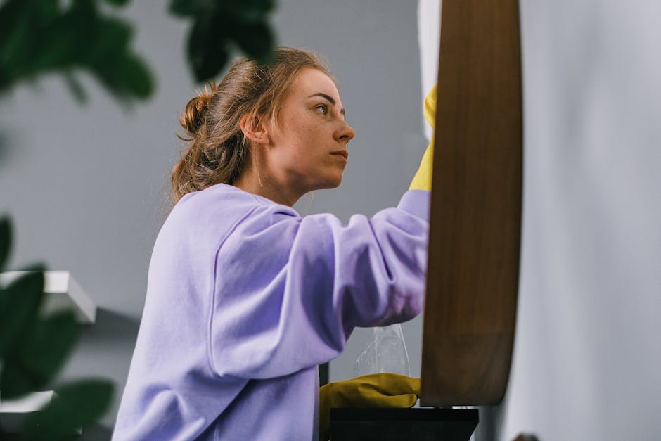 A close-up view of a person wearing bright blue rubber gloves performing surface cleaning on a white table or countertop, using a white cloth and a spray bottle with a green and white label. The scene